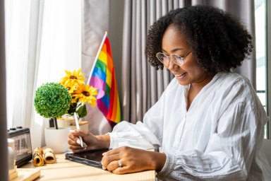African American girl is working at home with LGBTQ rainbow flag in her table for coming out of closet and pride month celebration to promote sexual diversity and equality in homosexual orientation