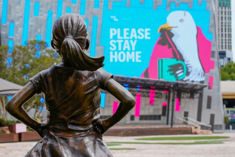 Fearless Girl statue looks at a “Please Stay Home” sign