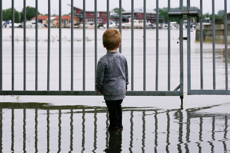 A child looks on as water floods through a fence,