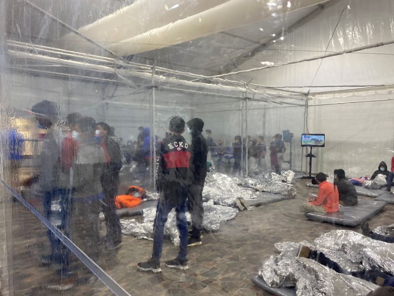 Migrants crowd a room with walls of plastic sheeting at