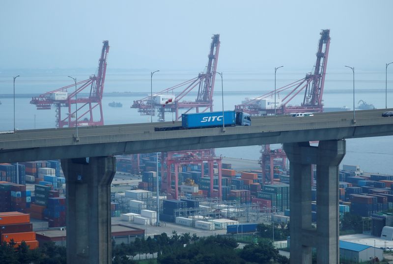 A truck carrying a shipping container travels past cranes at