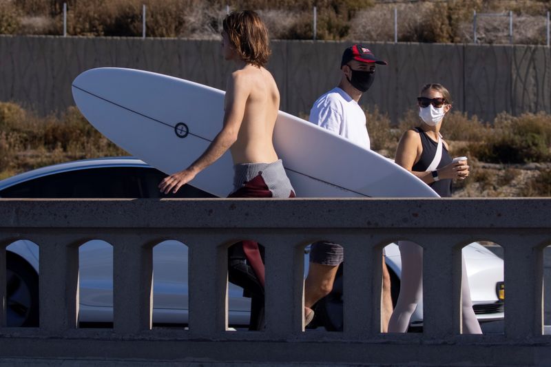 A surfer walks past masked pedestrians at the beach in