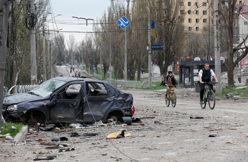 People ride bicycles near a destroyed car in Mariupol