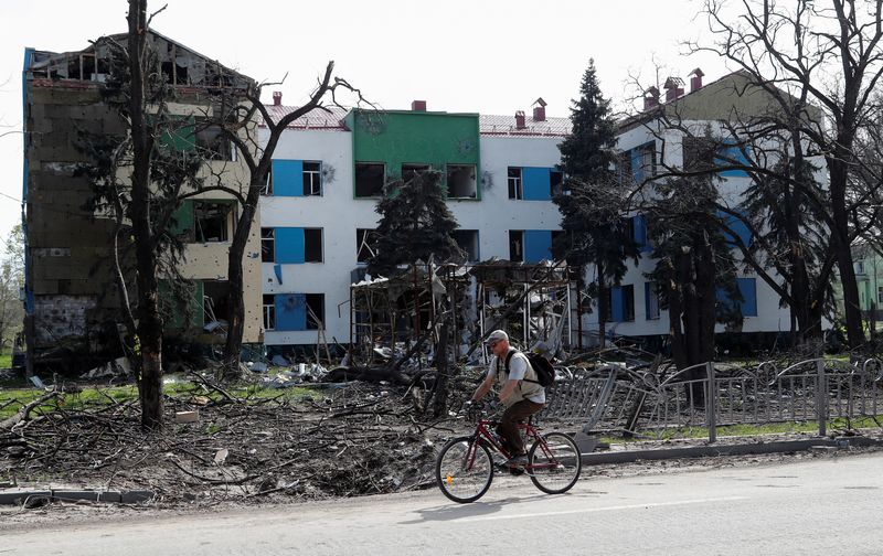 A man rides a bicycle near a damaged building in